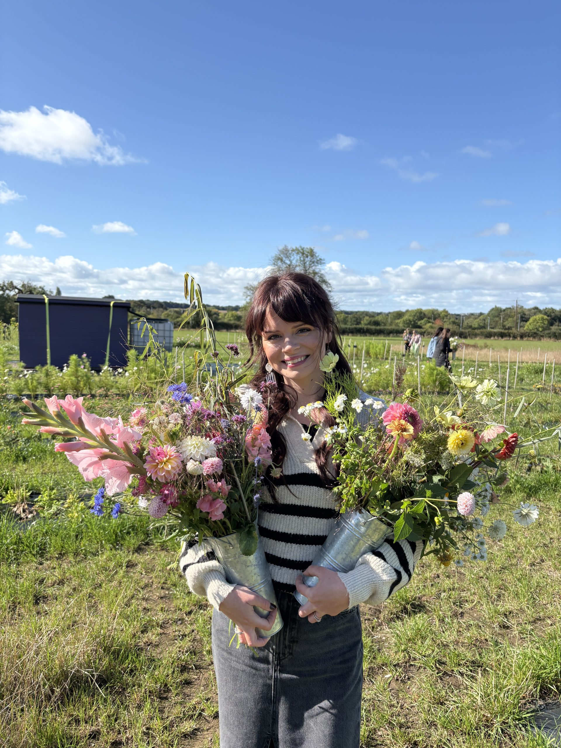 May & June founder and florist is smiling to camera with a flower farm her. She is carrying a bucket of handpicked British flowers in each arm